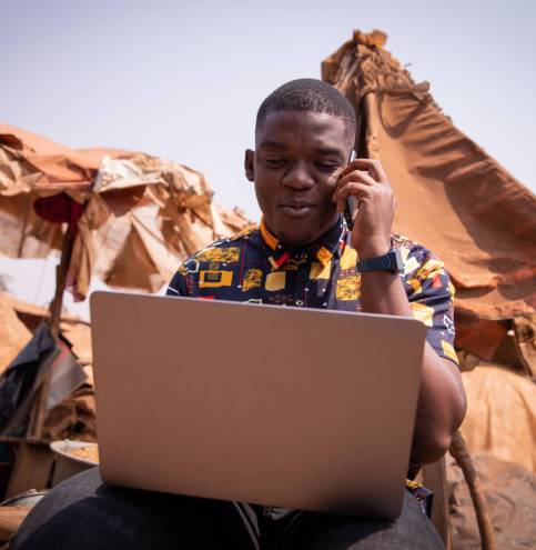 Young African boy works with his laptop and makes a phone call, he is at the market Young African boy works with his laptop and makes a phone call, he is at the market